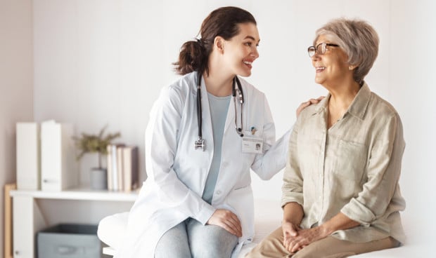 Doctora joven sonriendo y conversando con una paciente mayor durante una consulta médica en un entorno sanitario cercano y profesional.
