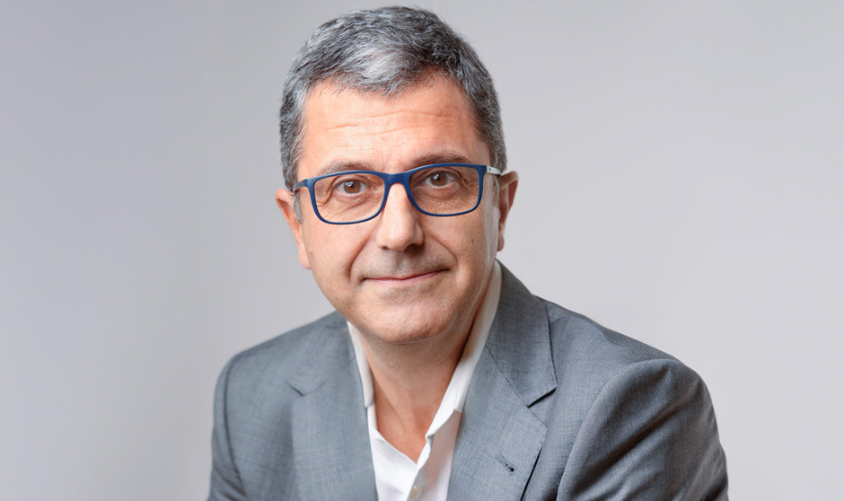 Un directivo empresarial con traje gris, gafas y sin corbata mira a cámara