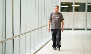 Persona con esclerosis lateral amiotrófica (ELA) caminando en un pasillo acristalado, con camiseta de apoyo a la concienciación sobre la ELA.