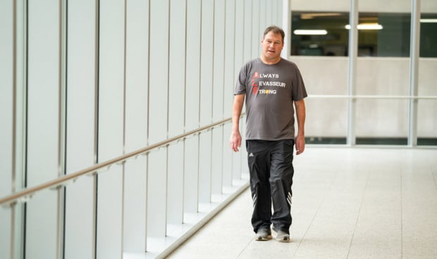 Persona con esclerosis lateral amiotrófica (ELA) caminando en un pasillo acristalado, con camiseta de apoyo a la concienciación sobre la ELA.