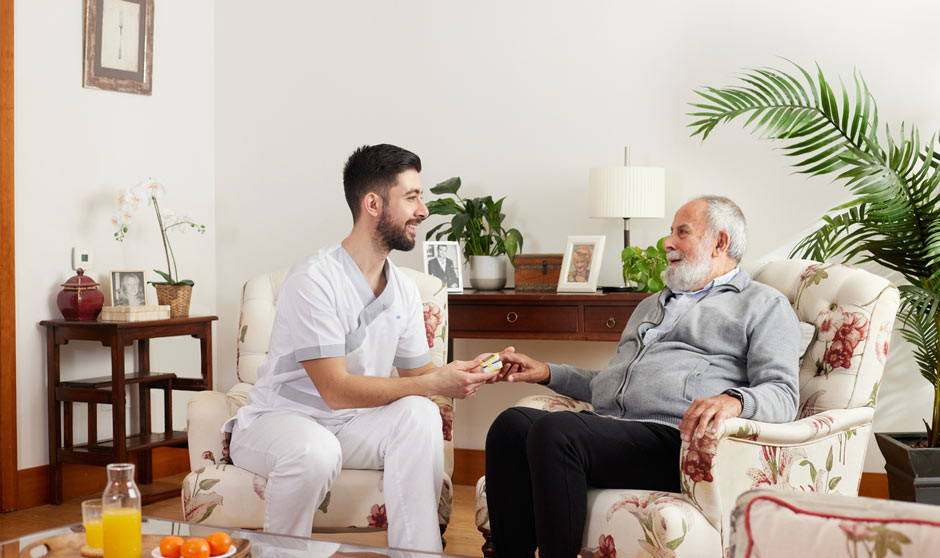 Profesional sanitario de Sanitas conversa y entrega medicación a un hombre mayor en su hogar, fomentando el cuidado personalizado y la autonomía.